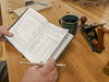 Hands holding a woodworking shop journal with pencil, smoothing plane, and coffee cup on workbench