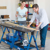 Two woodworkers measuring and marking a long board on a Kreg workbench with a circular saw below.