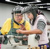Experienced woodturner instructing a student using a lathe with face shields in a workshop setting