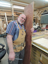 Woodworker holding a tall plywood panel in a workshop with tools and lumber around.