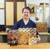 Woman in workshop displaying woodburned decorative plate, paddle, and patterned panel on workbench