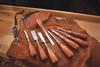 Set of eight bevel edge socket chisels with wooden handles arranged on a leather tool roll on a wooden workbench.