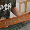 Close-up of a wooden sign engraved with the word Woodcraft, showing a smooth resin-filled design. The background shows