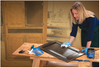 Woman applying wood stain to a cabinet door on a woodworking bench in a workshop.