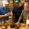 Three men examining wooden marble maze pieces on a workshop table with woodworking tools in background