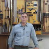 Woodworker standing in front of a wall-mounted tool cabinet with squares, planes, and clamps in a workshop.