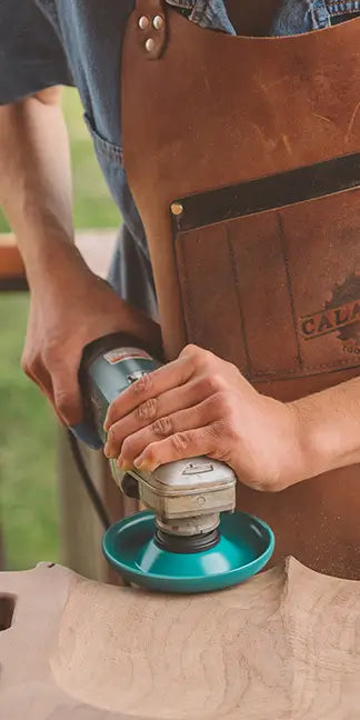 Person using a power sander on a piece of wood, wearing a leather apron.